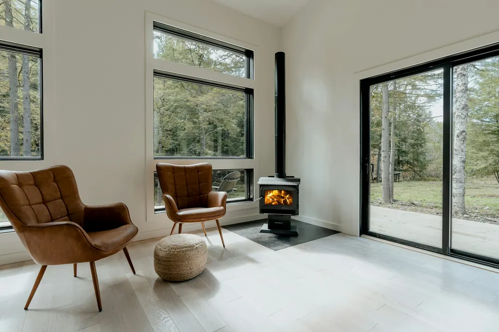 Deux fauteuils en cuir tournés vers un poêle à bois allumé, dans une pièce blanche ouverte sur le jardin